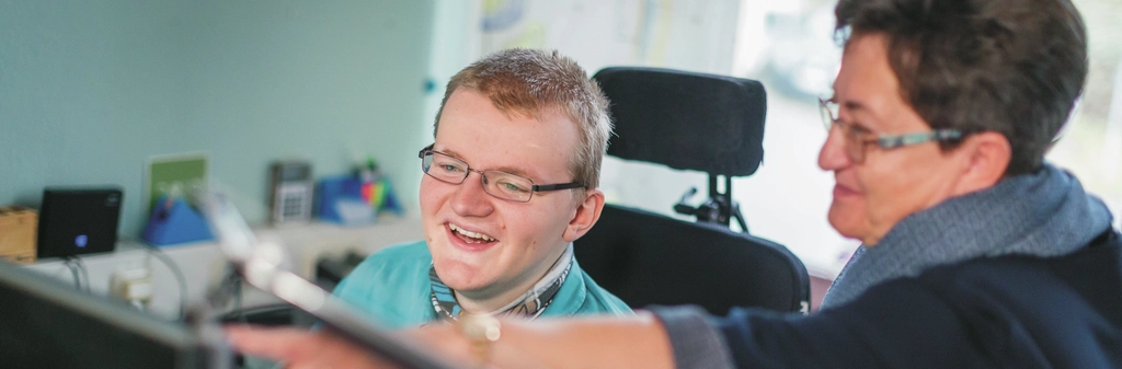 Ein junger Mann im Rollstuhl arbeitet am PC, eine Frau steht neben ihm und deutet auf den Bildschirm