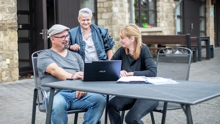 Zwei Personen sitzen draußen vor einem Laptop, eine dritte Person steht dabei.