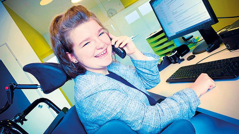 Eine junge Frau mit hellblauer Jacke sitzt in einem Büro vor einem Computer. Sie telefoniert.