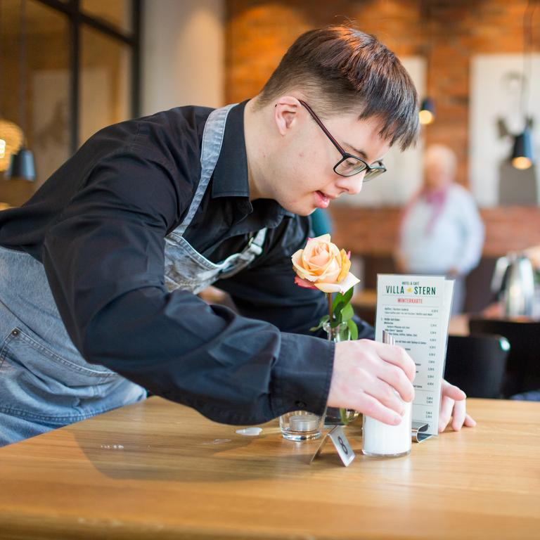 Ein junger Mann mit Downsyndrom steht über einen Cafetisch gebeugt. Er trägt eine Schürze und sortiert Speisekarte und Zuckerstreuer.