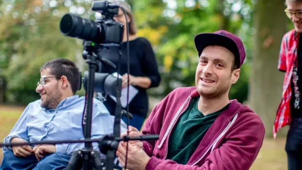 Zwei junge Männer im Rollstuhl nebn einer Kamera.