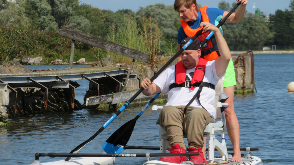 Auf einem Standup-Paddle, auf dem Wsser, sind ein Mann der auf einem Stuhl sitzt, sowie ein stehende Frau zu sehen. Beide halten ein Paddel in der Hand.