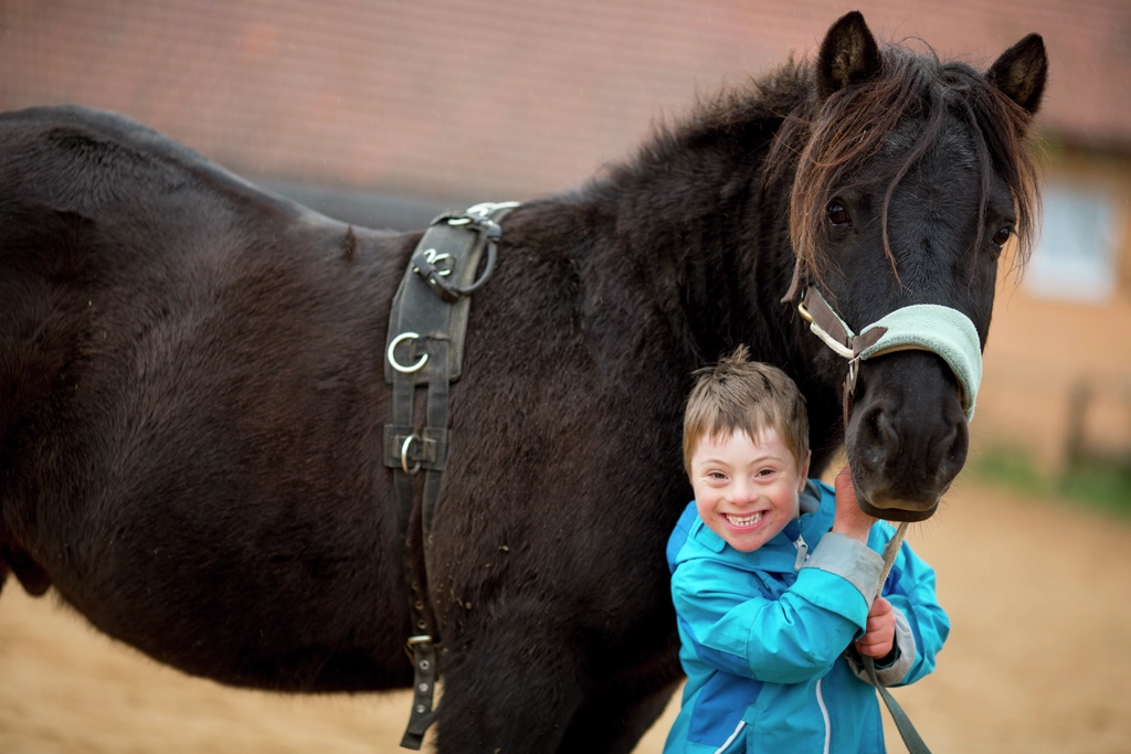 Ein Junge mit Down-Syndrom ist mit einem Pferd zu sehen, er lächelt in die Kamera. 