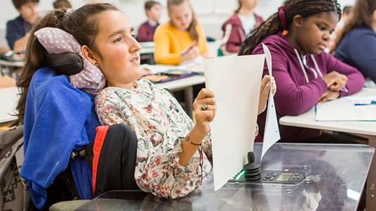 Eine Schülerin im Rollstuhl in einem gefüllten Klassenzimmer.