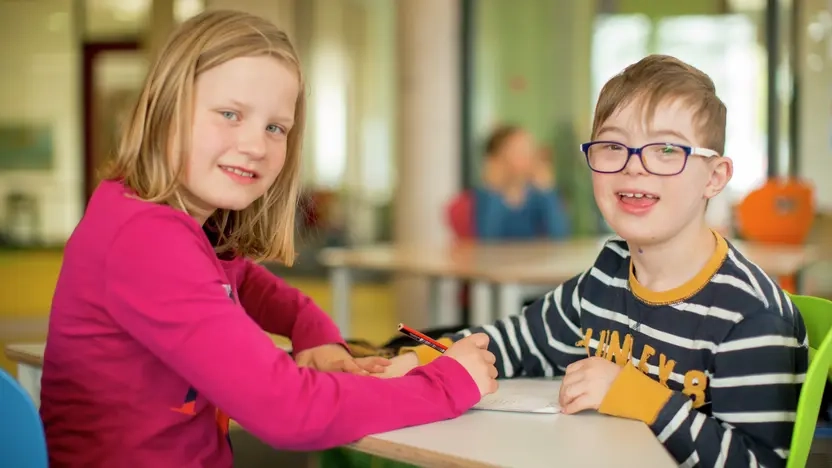Zwei Kinder sitzen sich am Tisch gegenüber und schreiben auf das gleiche Blatt Papier. 