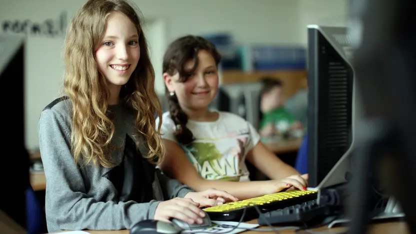 Zwei Mädchen sitzen vor einem Rechner. Beide lächeln in die Kamera.