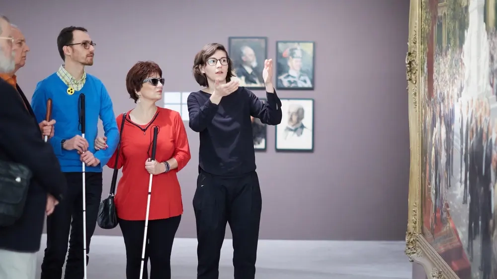Im Museum: Eine Frau erzählt verschiedenen Menschen mit Blindenstock etwas über ein Gemälde.