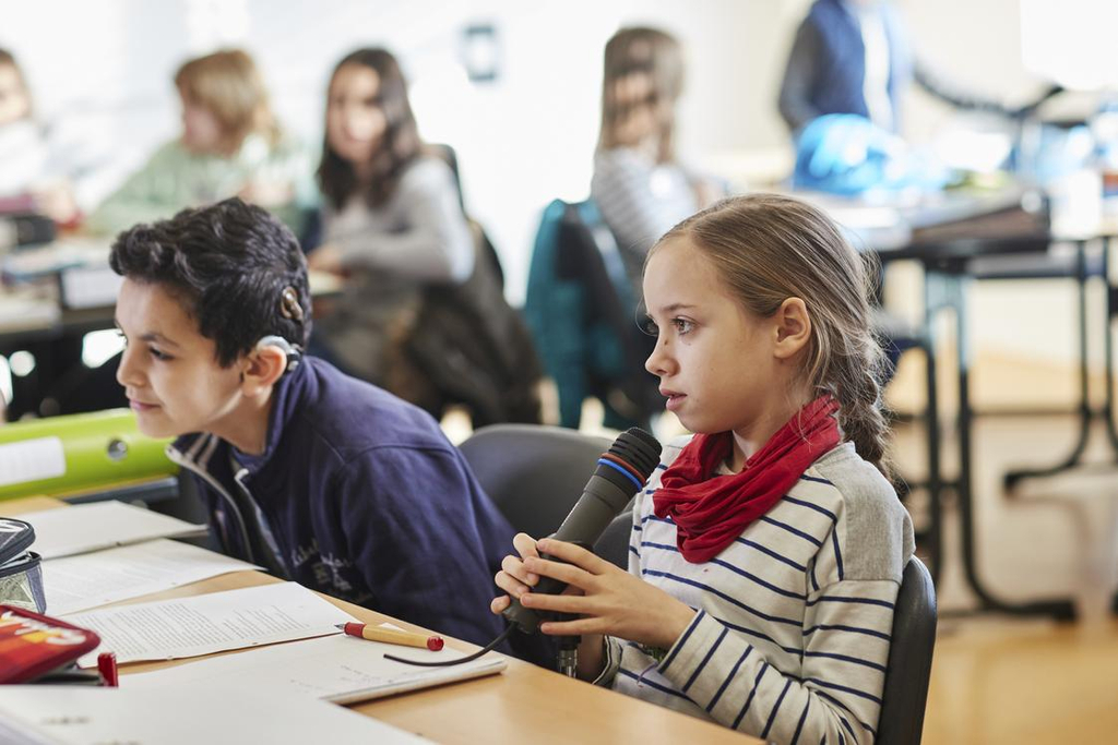 Ein Junge und ein Mädchen sitzen in einer Klasse. Das Mädchen spricht in ein Mikrofon.