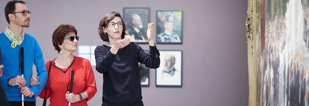 Im Museum: Eine Frau erzählt verschiedenen Menschen mit Blindenstock etwas über ein Gemälde.