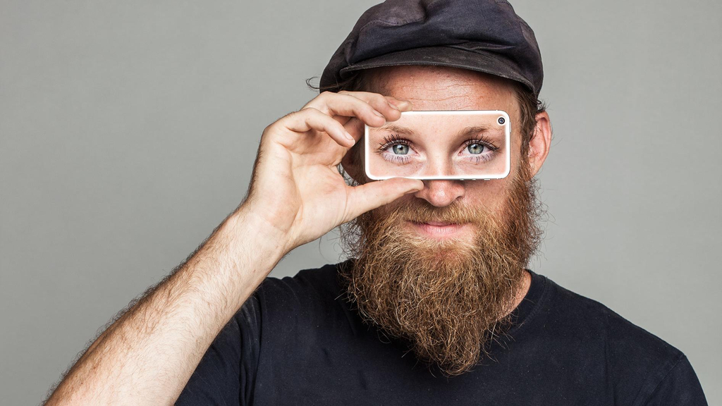 Junger Mann mit Schiebermütze und Rauschebart hält sich lächelnd ein Handy vor beide Augen, auf dem Display sind Augen eines anderen zu sehen