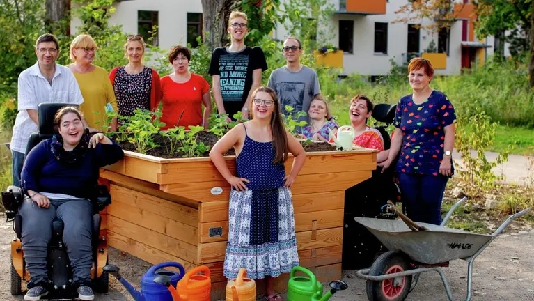 Die Bewohner der Wohngemeinschaft haben sich an einem Hochbeet versammelt. Drei Bewohnerinnen sitzen im Rollstuhl.