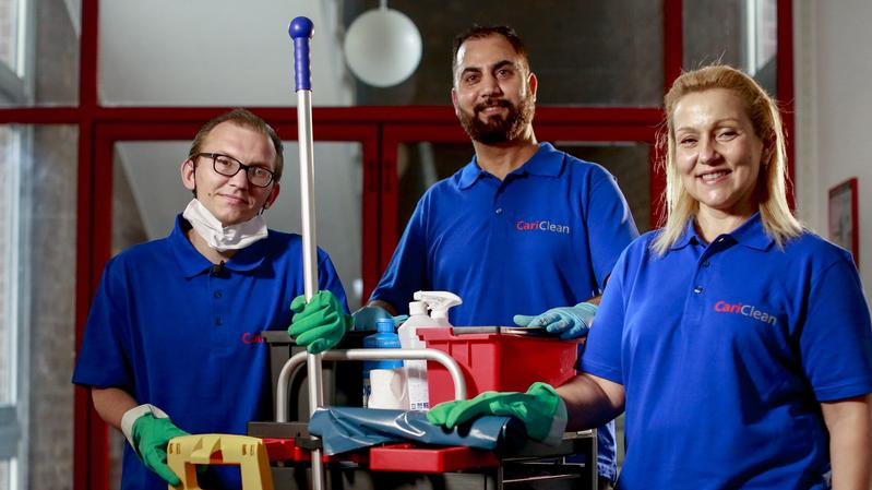 Zwei Männer und eine Frau stehen in Arbeitskleidung neben einem Reinigungswagen und lachen in die Kamera.