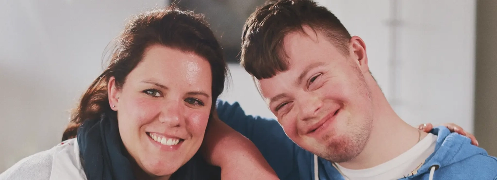 Eine Frau mit braunen Haaren und ein junger Mann mit Down-Syndrom haben einen Arm umeinander gelegt und lachen in die Kamera.