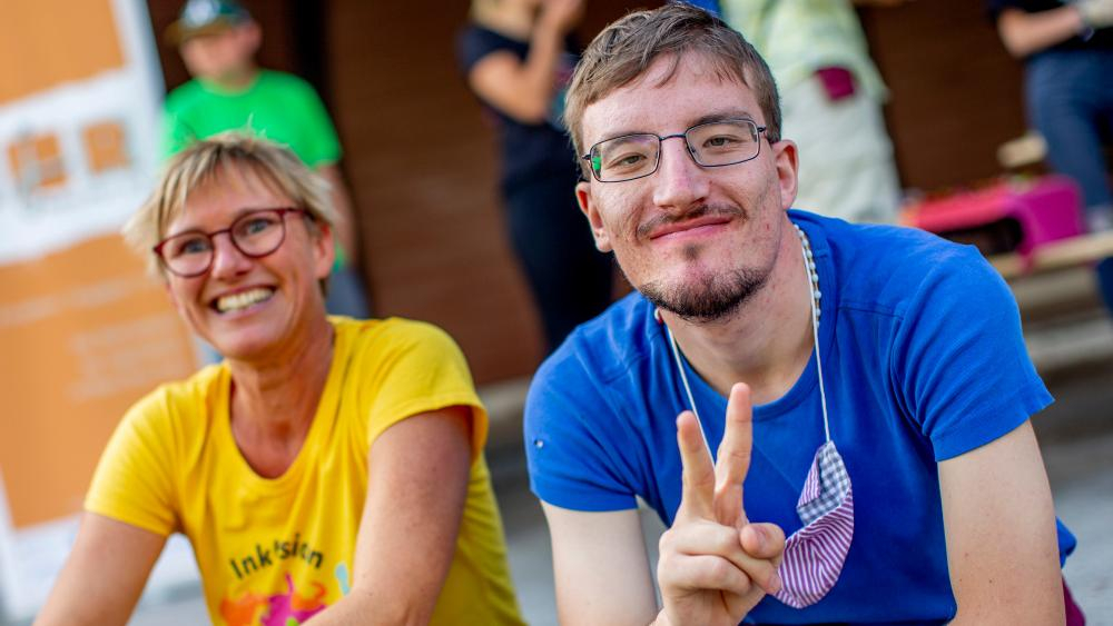 Eine Frau und ein Mann mit geistiger Behinderung sitzen lächelnd nebeneinander. er macht ein Victory-Zeichen