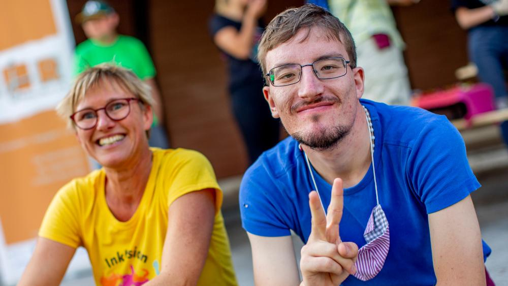 Eine Frau und ein Mann mit geistiger Behinderung sitzen lächelnd nebeneinander. er macht ein Victory-Zeichen