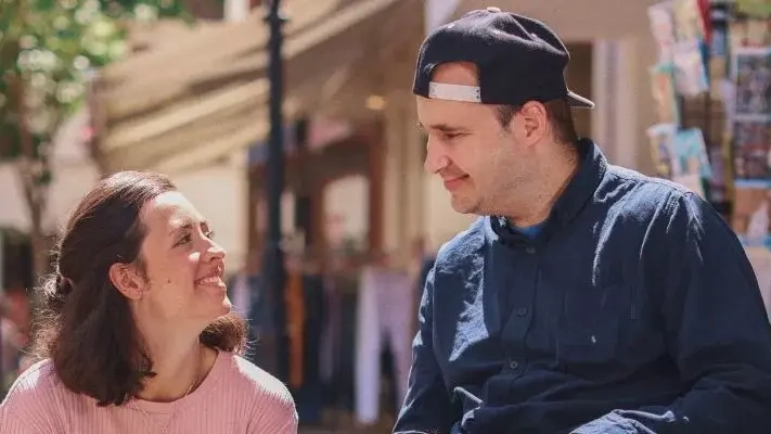 Ein Mann im Rollstuhl sitzt vor einer Buchhandlung. Neben ihm sitzt eine Frau in der Hocke. Die beiden lächeln sich an.