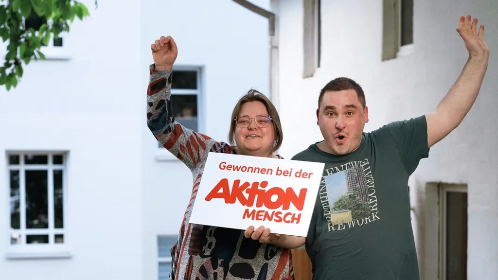 Ein Mann und Eine Frau stehen nebeneinander. Sie halten ein Schild auf dem steht: Gewonnen bei der Aktion Mensch. Bei reißen jubelnd die Arme in die Luft.