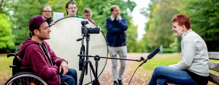 Ein junger Mann im Rollstuhl interviewt eine Frau in einem Park. Eine Kamera mit Stativ und Mikrofon sind auf die Frau gerichtet.