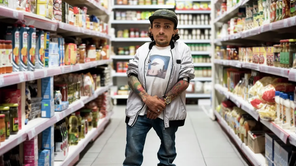 Ein Portraitbild von Gia Abramyan. Er steht im Gang eines Supermarkts zwischen zwei Regalreihen. Er hält die Hände vor dem Körper verschränkt und schaut ernst in die Kamera.