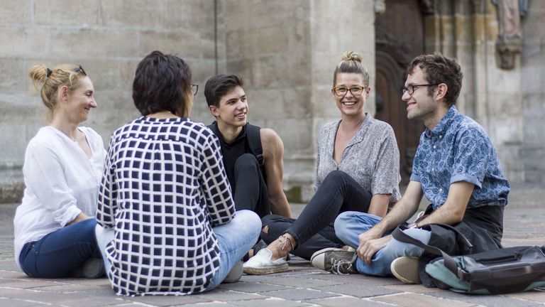 Ein Gruppe von Menschen sitzt vor einem Gebäude und sie unterhalten sich.