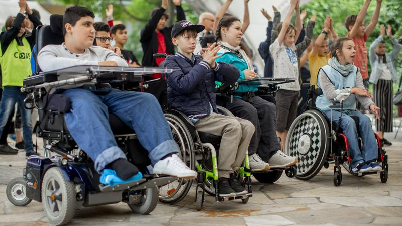 Mehrere Kinder sitzen und stehen in mehreren Reihen. Manche davon sitzen im Rollstuhl. Die meisten von ihnen jubeln.