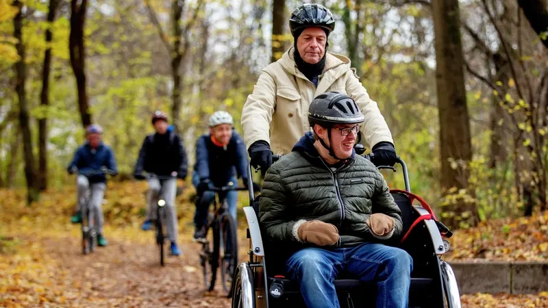 Im Vordergrund sind zwei Männer mit der Elektro-Rikscha zu sehen. Im Hintergrund folgen weitere Radfahrer auf dem Waldweg.