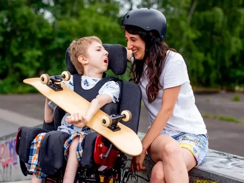 Ein Junge im E-Rolli steht auf einer Skate-Rampe. Er hält ein Skateboard im Arm. Neben ihm sitzt eine Frau mit Skate-Helm und lacht ihn an.