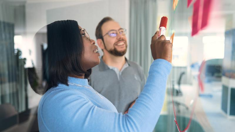 Eine Frau und ein Mann stehen gemeinsam vor einer Gläsernen Wand. Die Frau hält einen Stift in der Hand und zeigt damit auf einen pinken Klebezettel, der vor ihr an der Glasscheibe klebt.