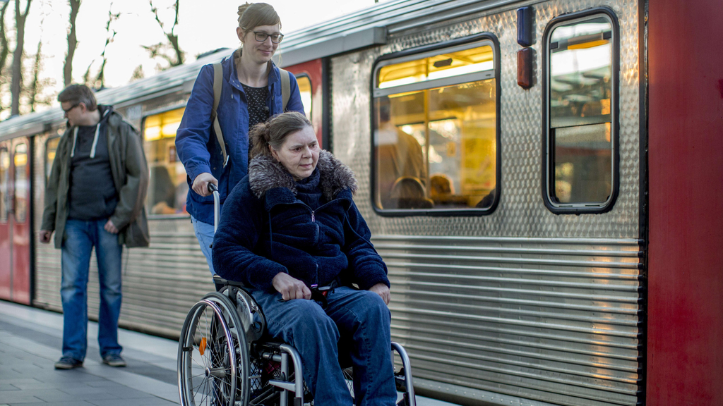 Eine ältere Frau sitzt in einem Rollstuhl, der von einer jüngeren Frau geschoben wird. Die beiden sind an einem Bahnsteig, an dem eine Bahn steht.