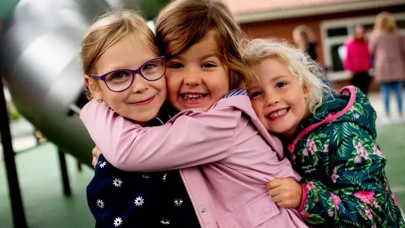 Drei Mädchen im Kindergarten umarmen sich herzlich und lachen in die Kamera.