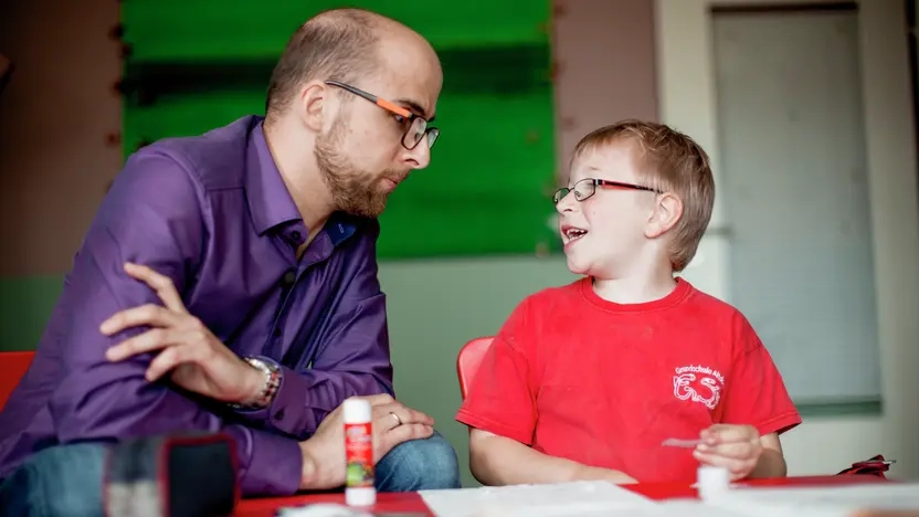 Ein Junge sitzt an einem Tisch und bastelt. Dabei wird er von einem jugen Mann beobachtet, der neben ihm sitzt. 