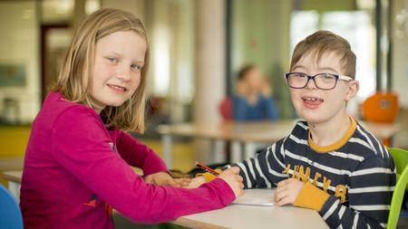 Zwei Kinder sitzen sich am Tisch gegenüber und schreiben auf das gleiche Blatt Papier. 