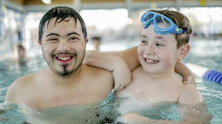Zwei Jugendliche sind zusammen in einem Schwimmbecken. Einer der beiden Jungen hat eine Behinderung, der andere nicht. Er trägt eine Schwimmbrille auf dem Kopf.
