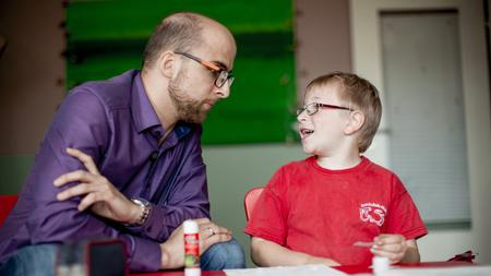 Ein kleiner Junge und ein Mann sitzen an einem Kindertisch und basteln gemeinsam. Der Junge trägt ein rotes T-Shirt, der Mann ein lilafarbenes Hemd. 