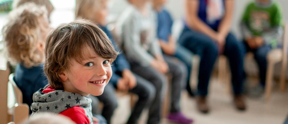 Ein Junge sitzt in einer Kindergartengruppe in einem Stuhlkreis und lächelt über die Schulter in die Kamera.