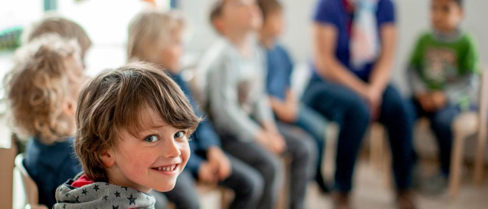 Ein Junge sitzt in einer Kindergartengruppe in einem Stuhlkreis und lächelt über die Schulter in die Kamera.