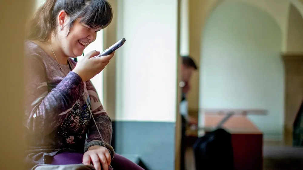 Carina sitzt an einer Fensterbank und spricht in ihr Handy. Dabei lächelt sie.