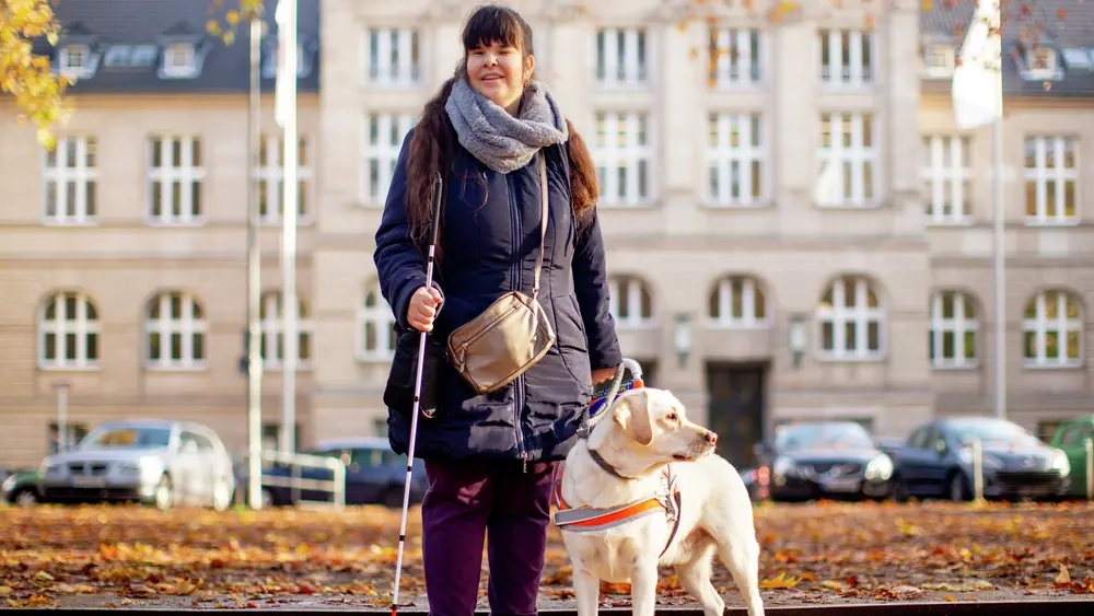 Carina steht mit ihrem Blindenstock und Hund Pitou vor einem weißen Gebäude und lächelt in die Kamera.