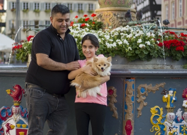 Ein Mädchen hält einen Hund auf dem Arm, der Vater steht daneben, sie stehen draußen vor einem bunt verziertem Brunnen