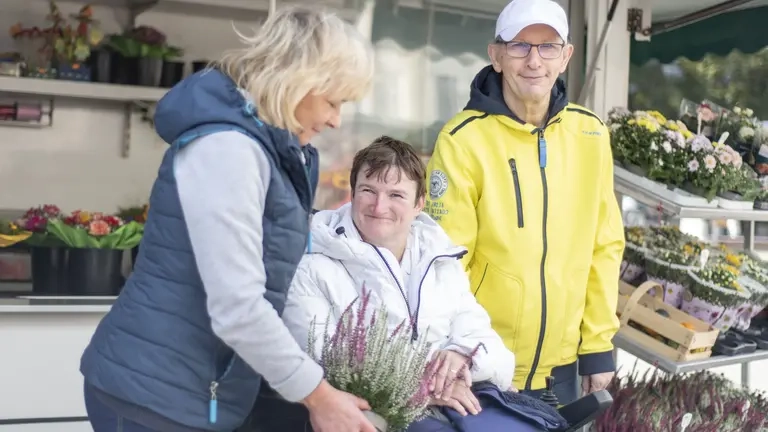 Drei Personen sind an einem Blumenstand auf einem Platz, eine Person sitzt im Rollstuhl