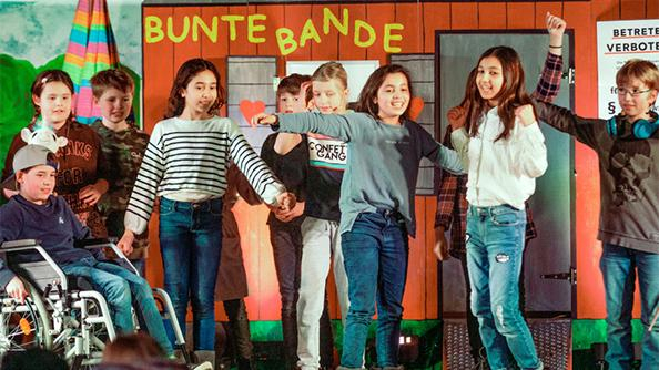 Kinder auf einer Theaterbühne, die eine Szene aus dem Bunte Bande-Musical präsentieren.