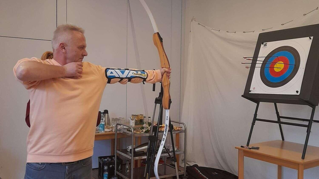 Ein Mann zielt mit einem Bogen auf eine Zielscheibe. Er steht in einem Raum.