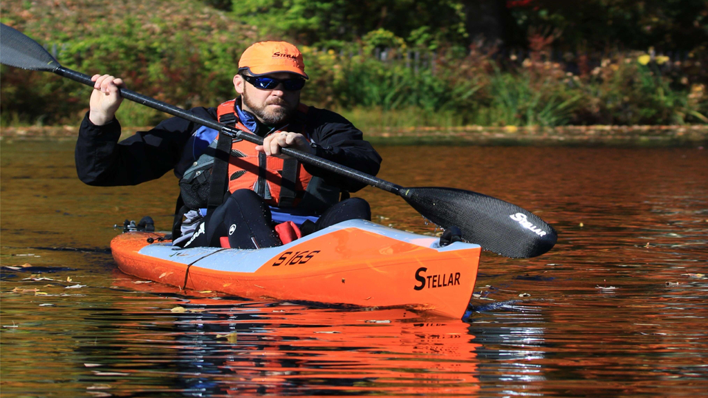 Ein Mann mit oranger Kappe und Sonnenbrille sitzt in einem orangen Kajak und hält ein Paddel in der Hand.