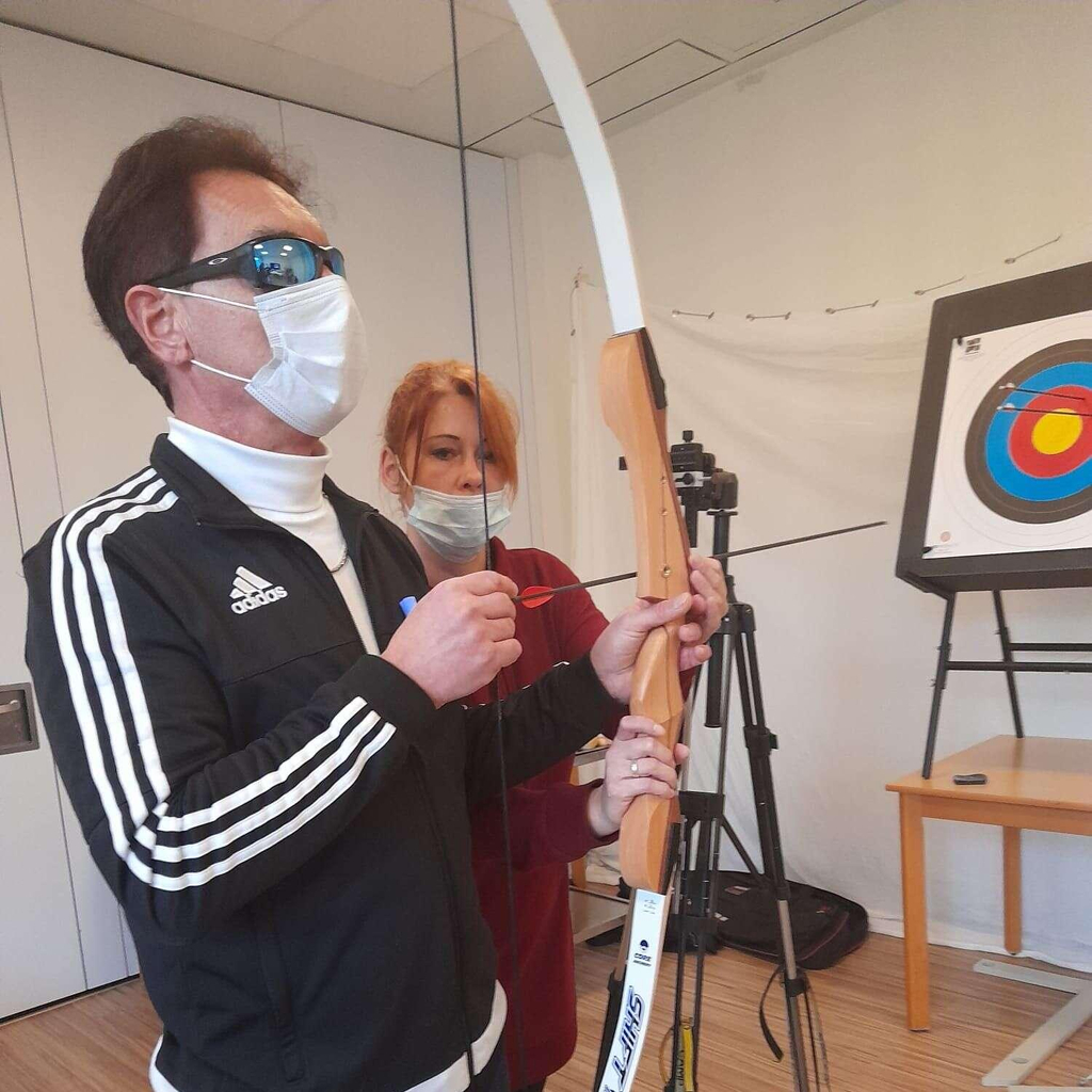 Ein Mann mit einer Sonnenbrille hält einen Bogen in der Hand. Eine Frau daneben hilft ihm, den Pfeil einzusetzen. Am Bildrand ist eine Zielscheibe zu sehen.
