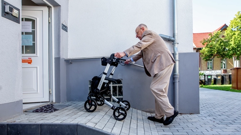 Ein älterer Mann geht mit einem Gehwagen über eine kleine Rampe in eine Behandlungspraxis.