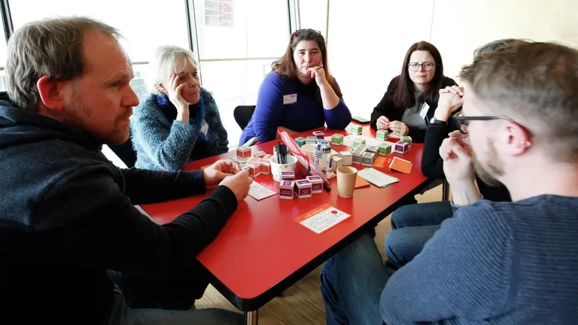 Mehrere Personen sitzen an einem Tisch und diskutieren. Vor ihnen liegen Arbeitsmaterialien auf dem Tisch.