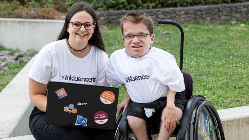 Max und Cristina sitzen in Inkluencer-T-Shirts mit einem Laptop im Freien und lächeln in die Kamera.