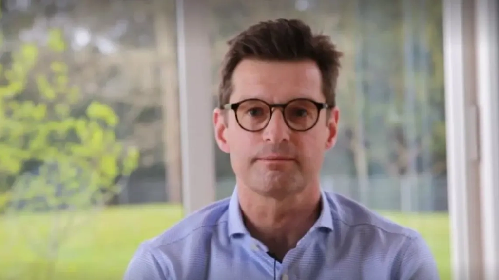 Ein Mann im mittleren Alter, mit brauen Haaren und dunkler Brille und blauem Hemd schaut in die Kamera.