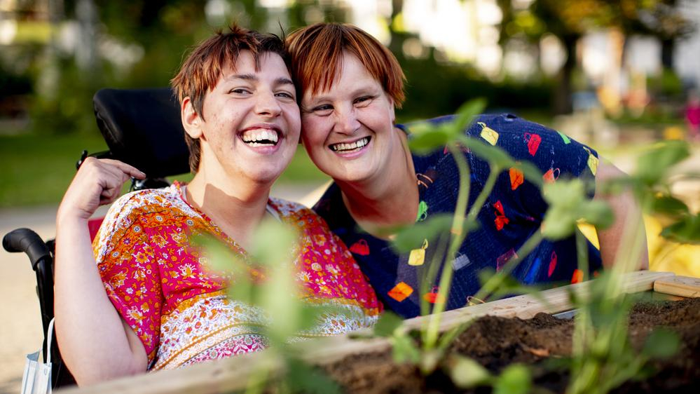 Zwei Frauen in bunt gemusterten Oberteilen sind hinter einem Hochbeet zu sehen. Eine der beiden Frauen sitzt in einem Rollstuhl. Sie stecken die Köpfe zusammen und lachen in die Kamera.