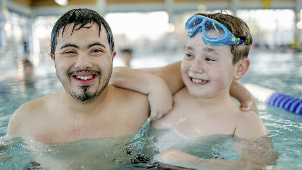 Zwei Jugendliche sind zusammen in einem Schwimmbecken. Einer der beiden Jungen hat eine Behinderung, der andere nicht. Er trägt eine Schwimmbrille auf dem Kopf.